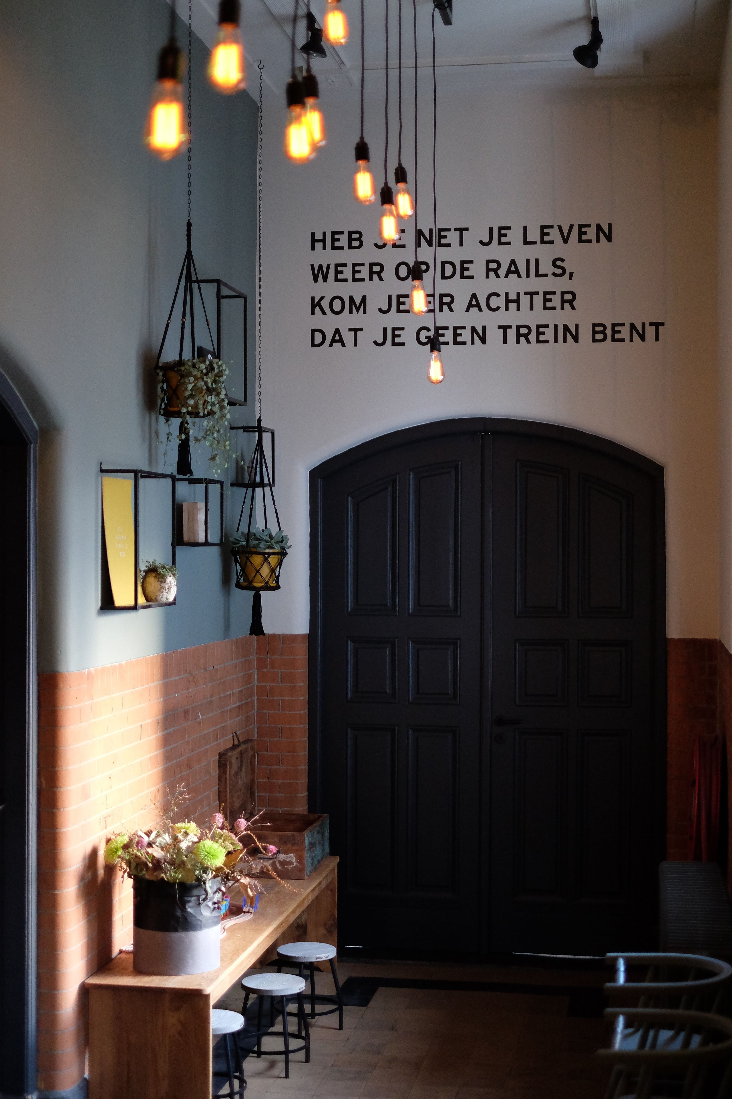 A hallway with hanging light bulbs, a black door, and a bench with stools.