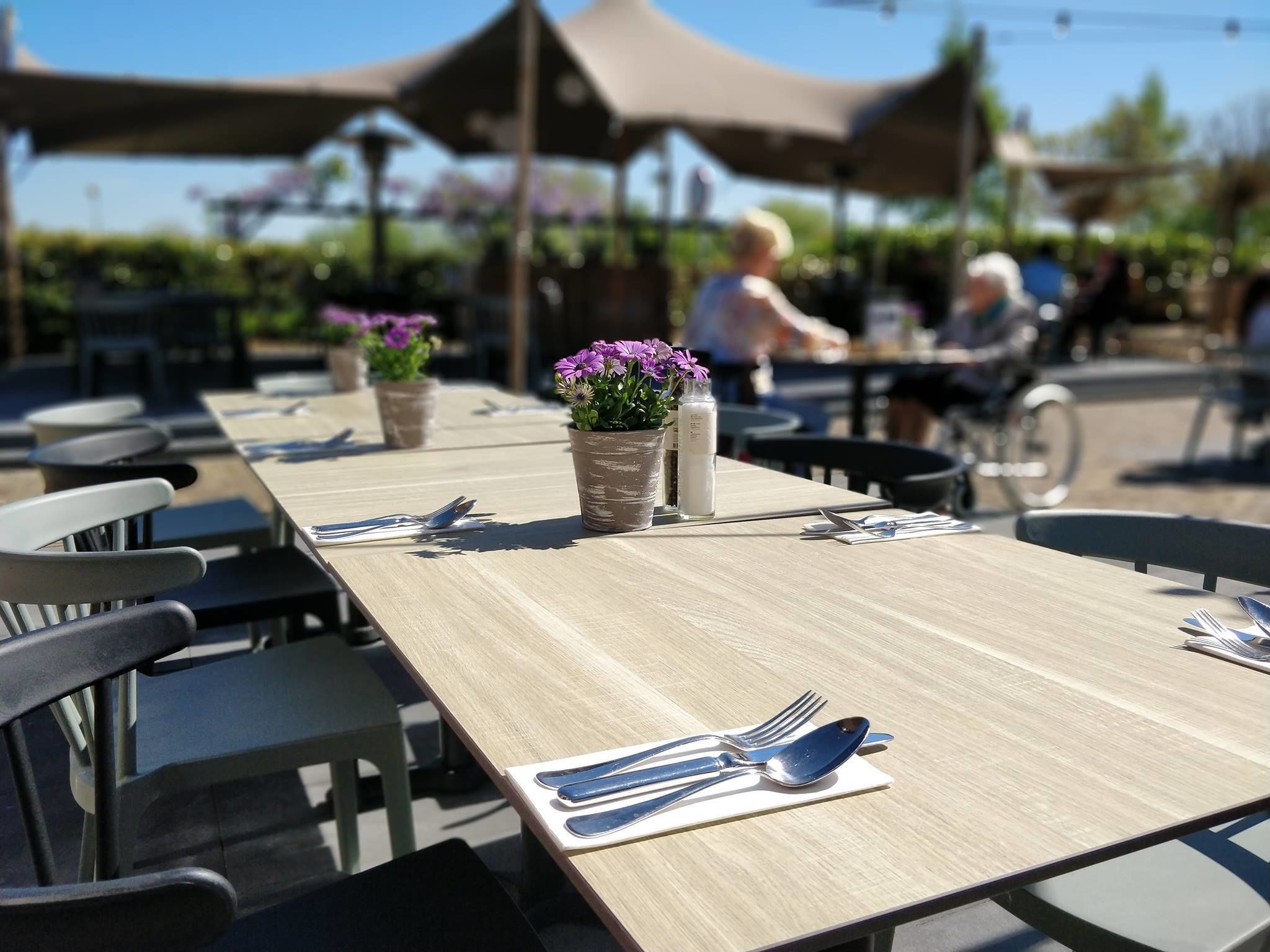 Un área de comedor al aire libre con mesas y sillas de madera, y macetas con flores moradas.