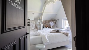 Een slaapkamer met een wit bed, een klein krukje, een witte ventilator en een bakstenen muur.