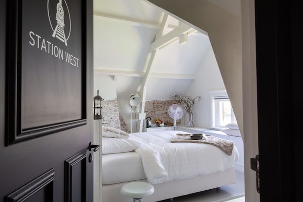 A bedroom with a white bed, a small stool, a white fan, and a brick wall. - Het Spoorhuis Uithoorn (Uithoorn)