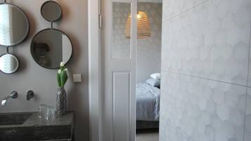 A modern bathroom with a geometric patterned wall, a round mirror, a light fixture, and a door leading to a bedroom.
