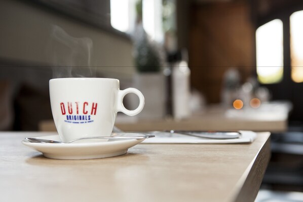 A white coffee cup with "DUTCH ORIGINALS" text on a saucer, steam rising from the cup, on a wooden table.