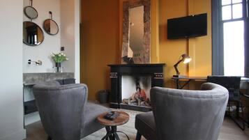 A living room with two grey armchairs, a small round table with a remote control, a fireplace with a stone surround, a flat-screen TV mounted on the wall, and a window providing natural light.
