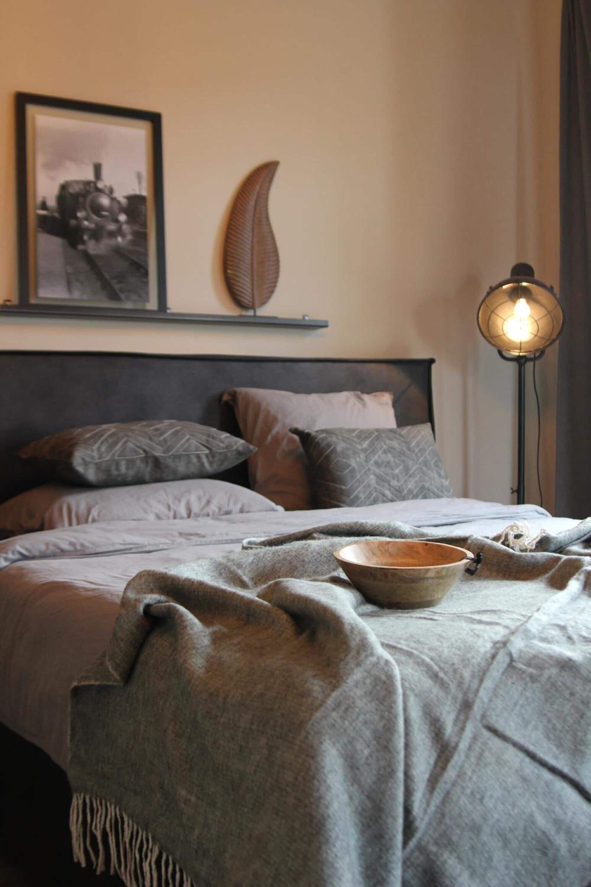 A neatly made bed with a gray blanket and pillows, a wooden bowl on the bed, a wall-mounted picture of a train, and a floor lamp with a light bulb.
