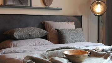 Een net opgemaakt bed met een grijze deken en kussens, een houten kom op het bed, een aan de muur hangend schilderij van een trein, en een vloerlamp met een gloeilamp.