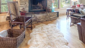 A stone fireplace with a mantel, a large window, and a view of a forest. There are two chairs and a sofa in the room.