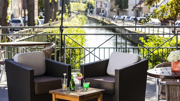 A terrace with wicker furniture, a table with drinks, and a view of a canal with buildings and parked cars.