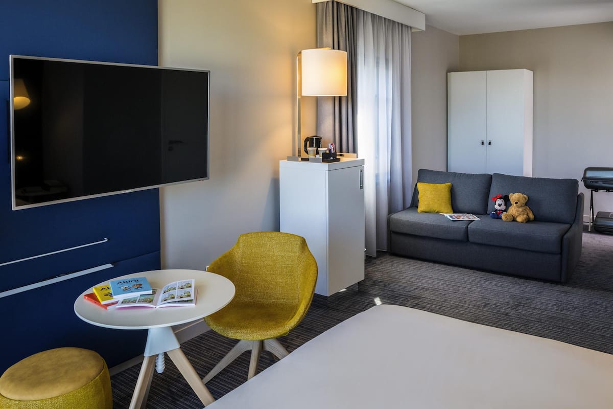 A hotel room with a TV, a sofa, a small table with books, a yellow chair, and a cabinet.