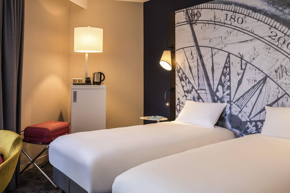A hotel room with two beds, a desk, a lamp, a small refrigerator, and a chair.