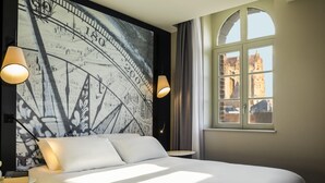 A hotel room with a large bed, two bedside lamps, a window with a view of buildings, and a wall mural.