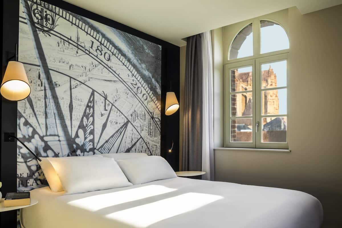 A hotel room with a large bed, two bedside lamps, a window with a view of buildings, and a wall mural.
