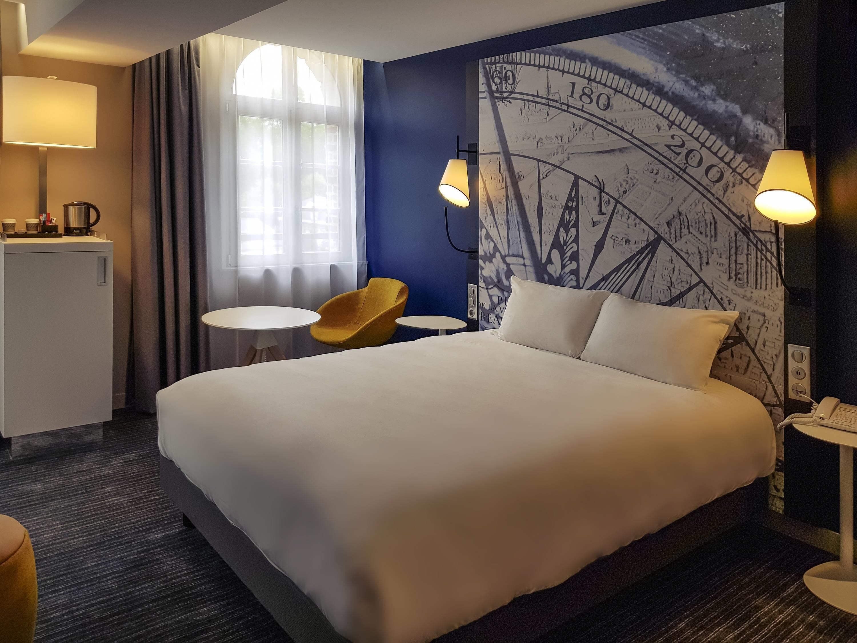 Hotellihuone, jossa on suuri sänky, kaksi yöpöytälamppua, pieni pöytä ja tuoli, kahvinkeitin sekä seinä, jossa on suuri seinämaalaus, joka esittää suurta kelloa.