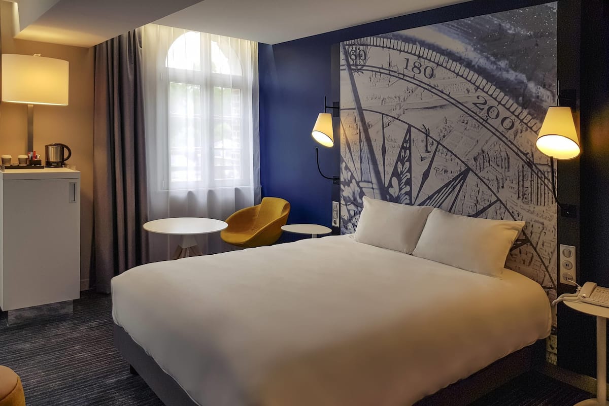 A hotel room with a large bed, two bedside lamps, a small table with a chair, a coffee machine, and a wall with a large clock mural.
