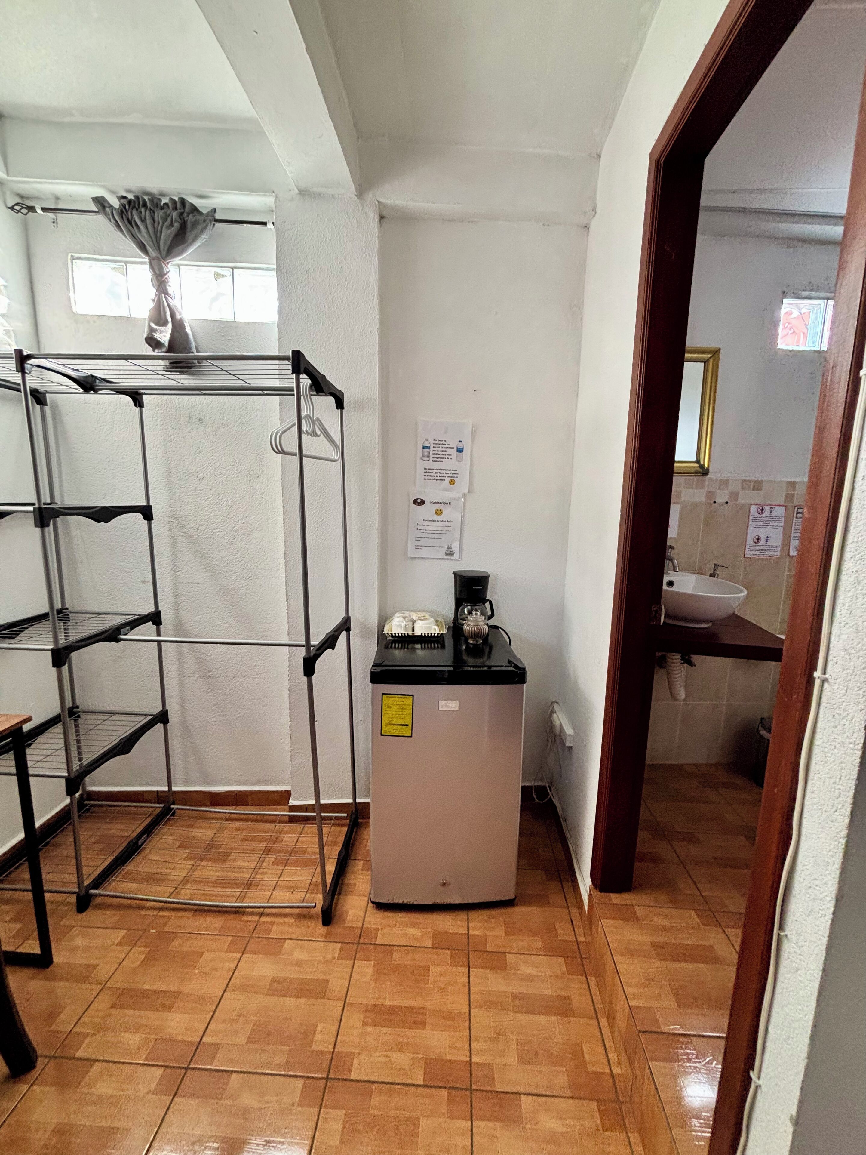 Eine kleine Küche mit Kühlschrank, Kaffeemaschine und Spüle. Ein Metallregal mit Ablagen sowie ein Fenster mit Vorhang sind vorhanden.