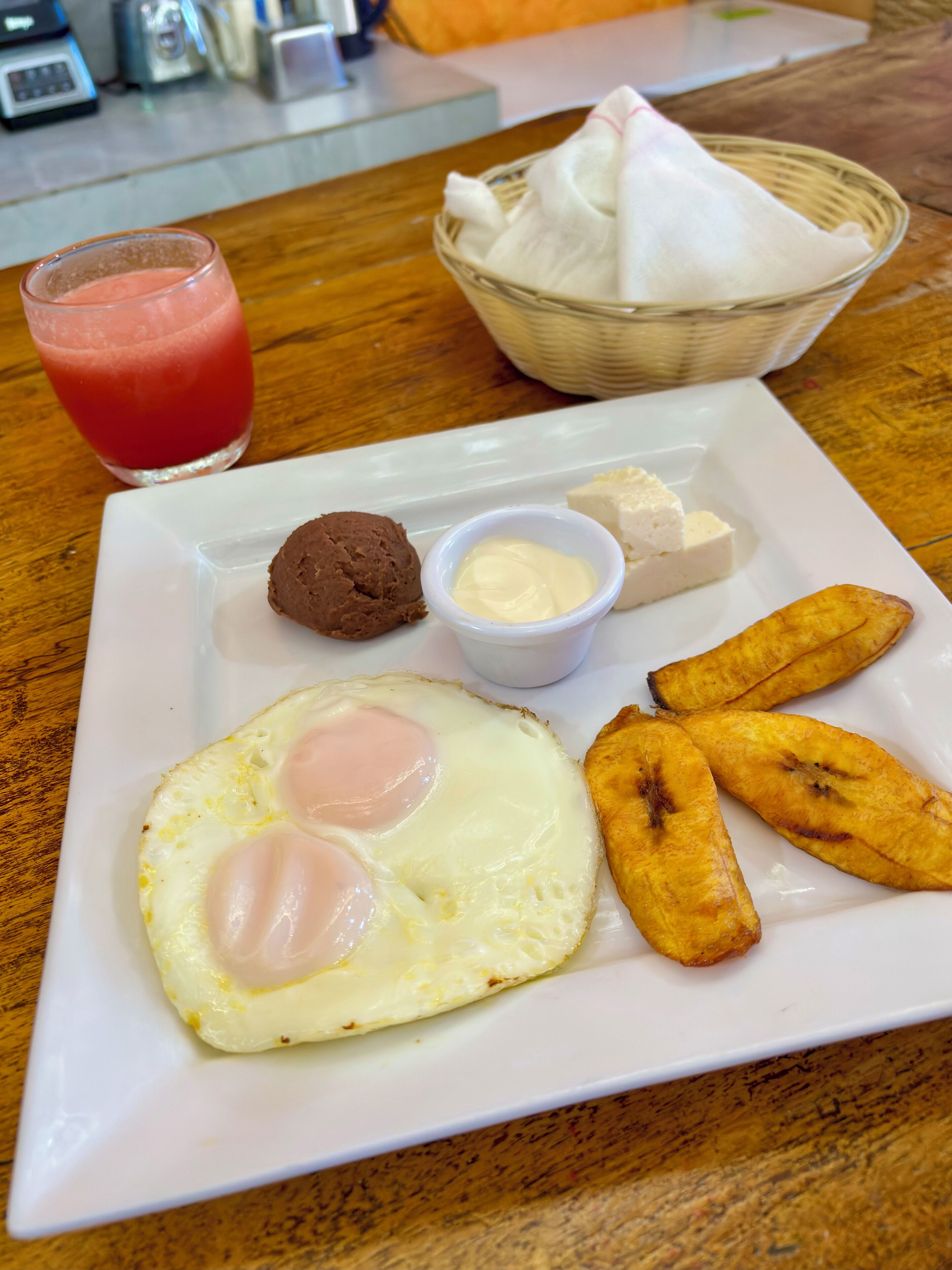 A breakfast plate with fried eggs, a scoop of chocolate ice cream, a small bowl of butter, a glass of red juice, and fried plantains on a wooden table.