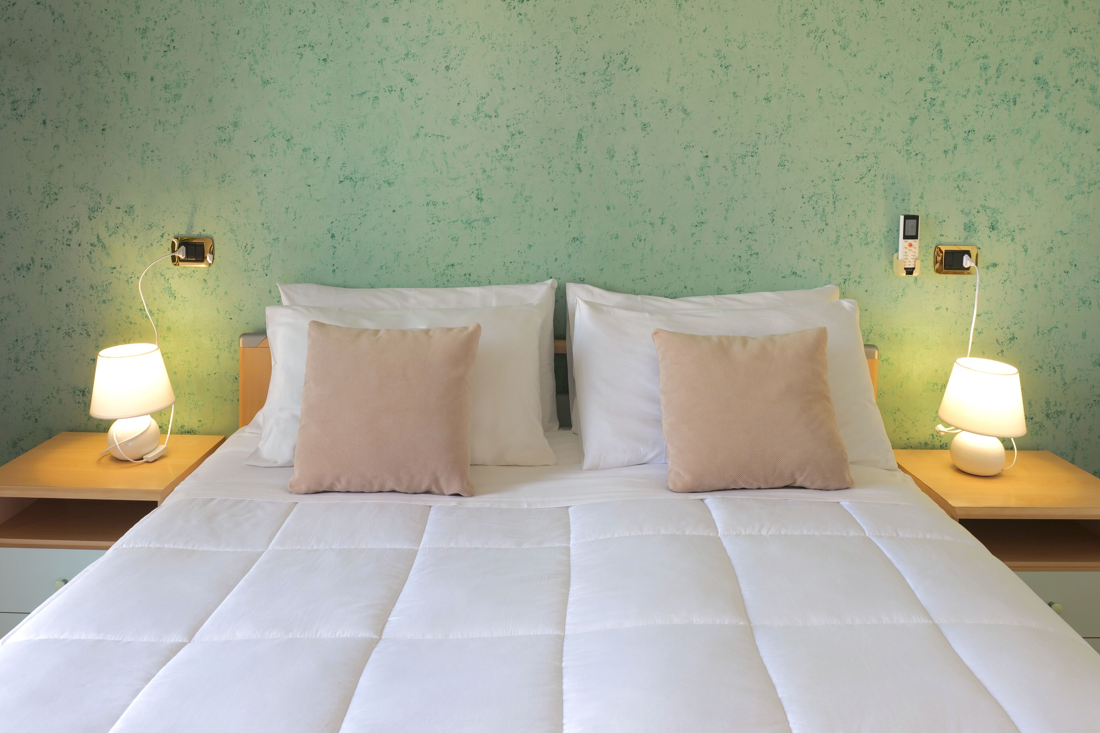 A neatly made bed with white bedding and two beige pillows, flanked by bedside tables with lamps, against a textured green wall.