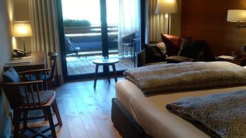 Habitación de hotel con piso de madera, cama con manta estampada, escritorio con lámpara, una silla y vista al exterior a través de una puerta de vidrio.