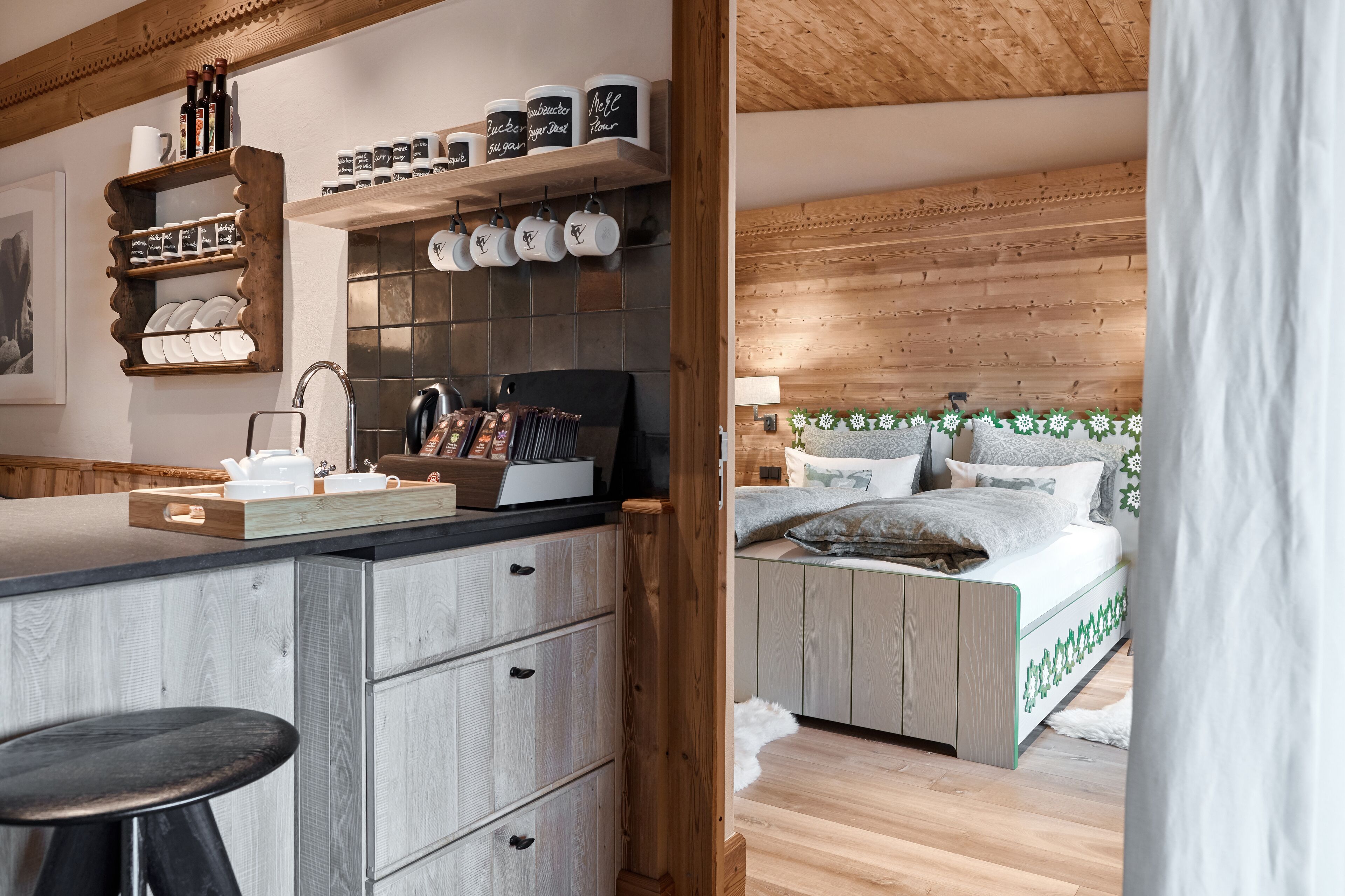 Eine moderne Küche mit Holzschränken, einer Arbeitsplatte und einem Barhocker. Ein Bett mit Holz Kopfteil und einem Zierkissen sind ebenfalls vorhanden.