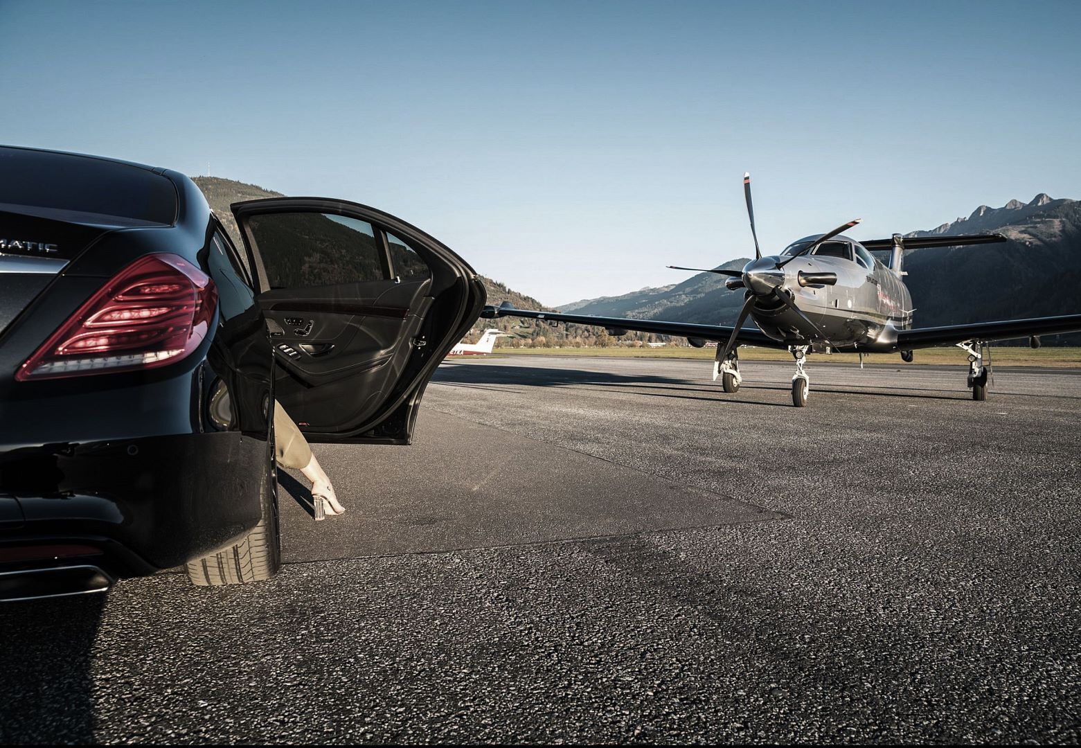 A parked car with its door open, an airplane on a runway, and mountains in the background.