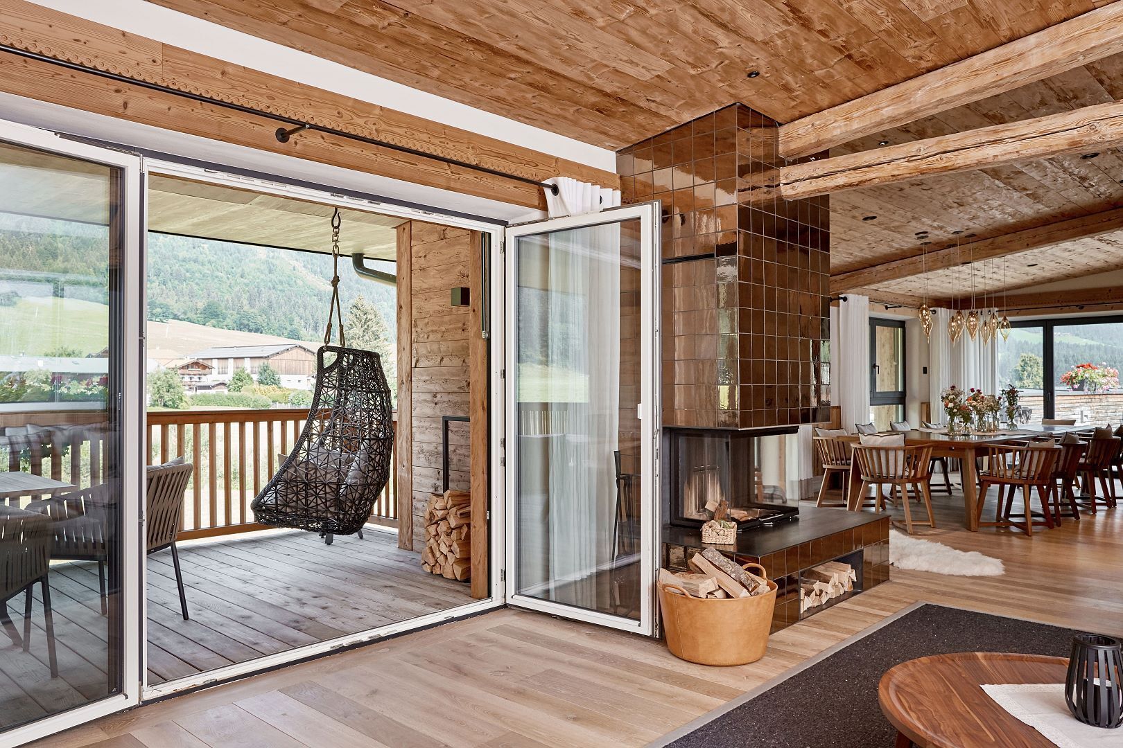 Ein geräumiger Wohnbereich mit Holzbalkendecke, einem Hängekorbstuhl, einem Kamin und einem Essbereich mit Tisch und Stühlen.