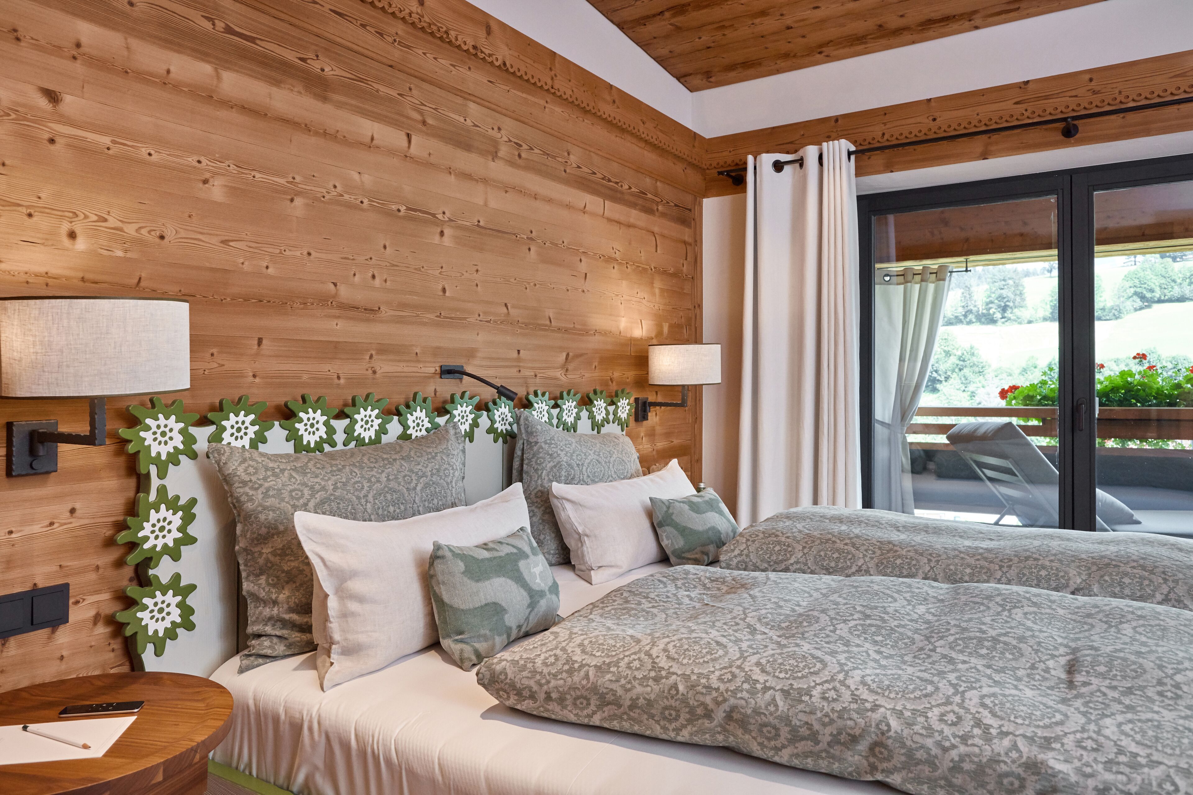 Ein Schlafzimmer mit Holzkopfteil, zwei Betten mit gemusterten Betttextilien, einem Nachttisch mit Lampe und Blick auf einen Garten durch eine Schiebeglastür.