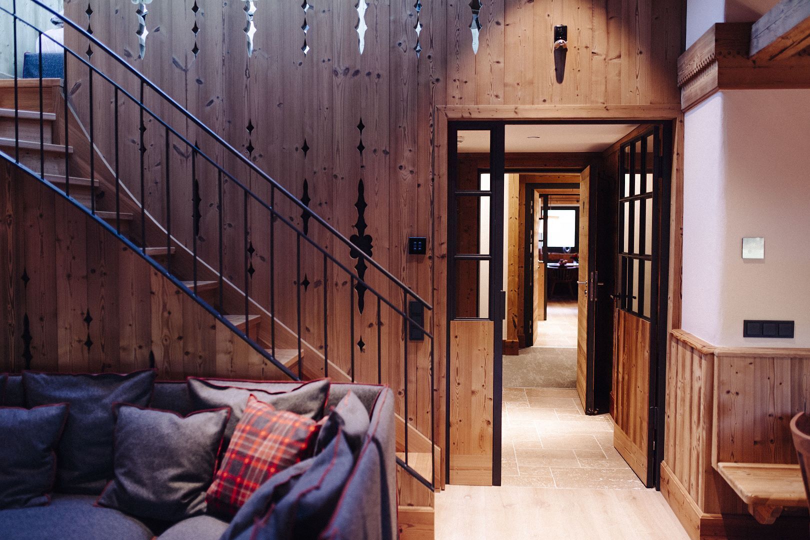 Un interior de madera con una escalera, un sofá con cojines escoceses y un pasillo que conduce a otra habitación.