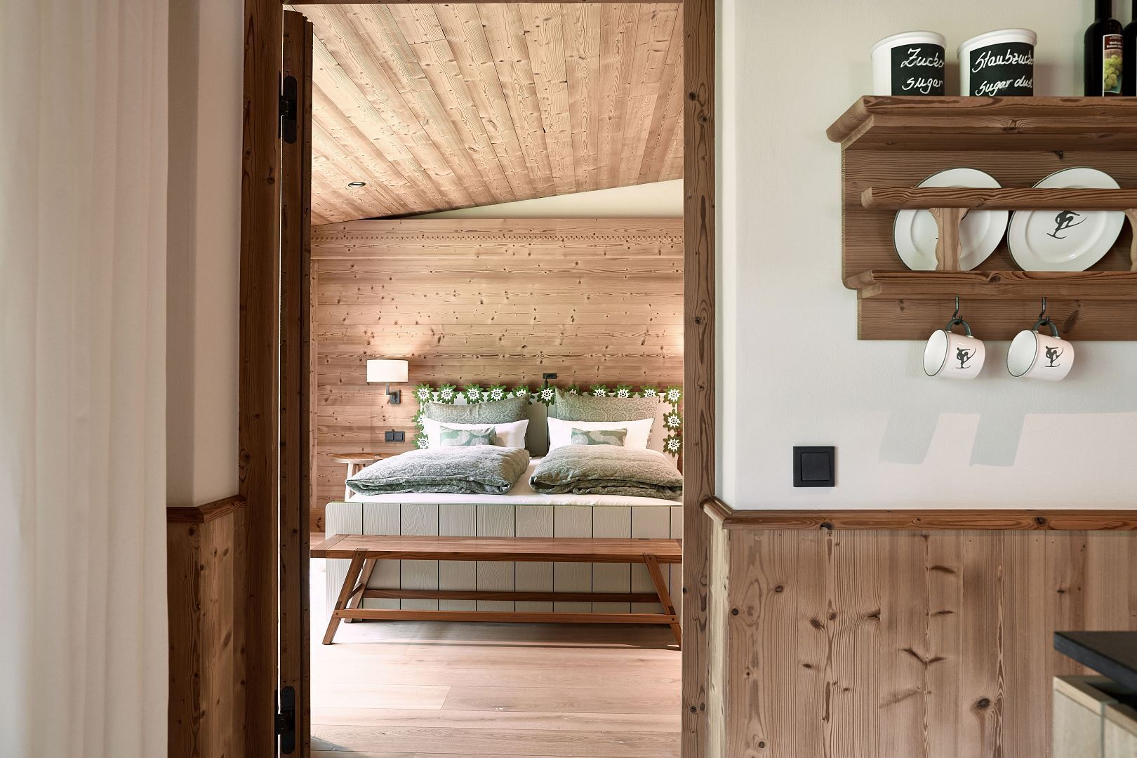 A wooden interior with a loft bed, a shelf with plates and mugs, and a wooden ceiling.