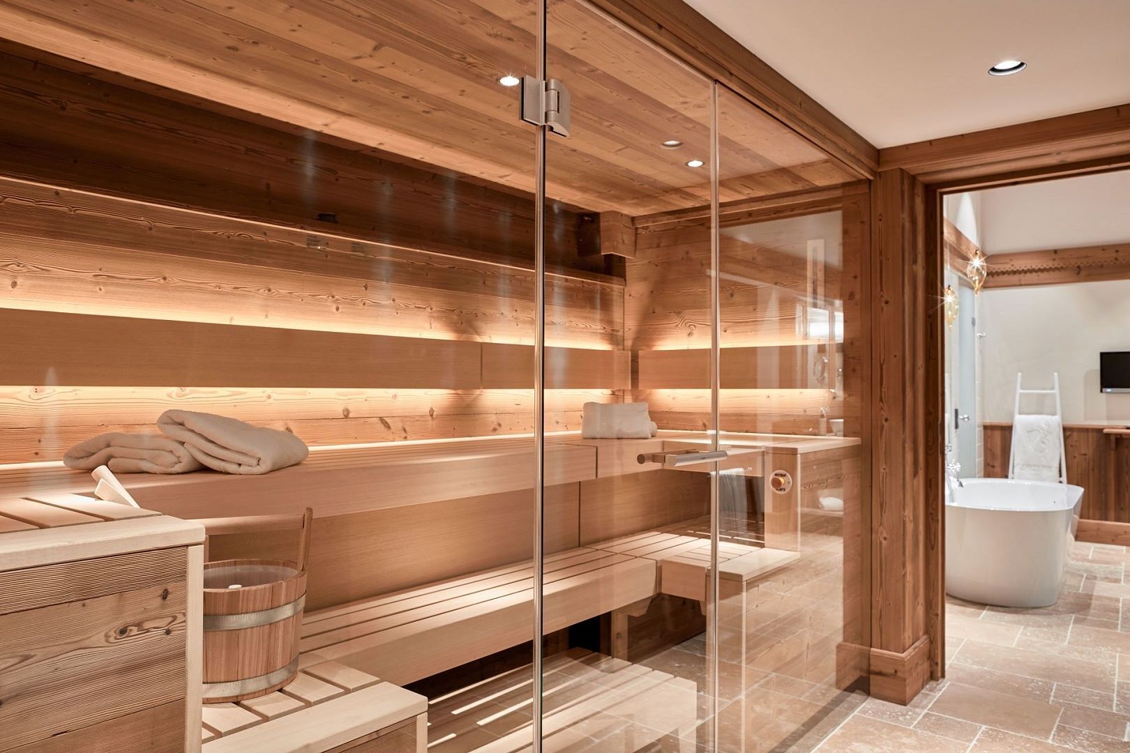 A modern spa room with wooden paneling, a glass-enclosed sauna, a wooden bench, and a stone-tiled floor.