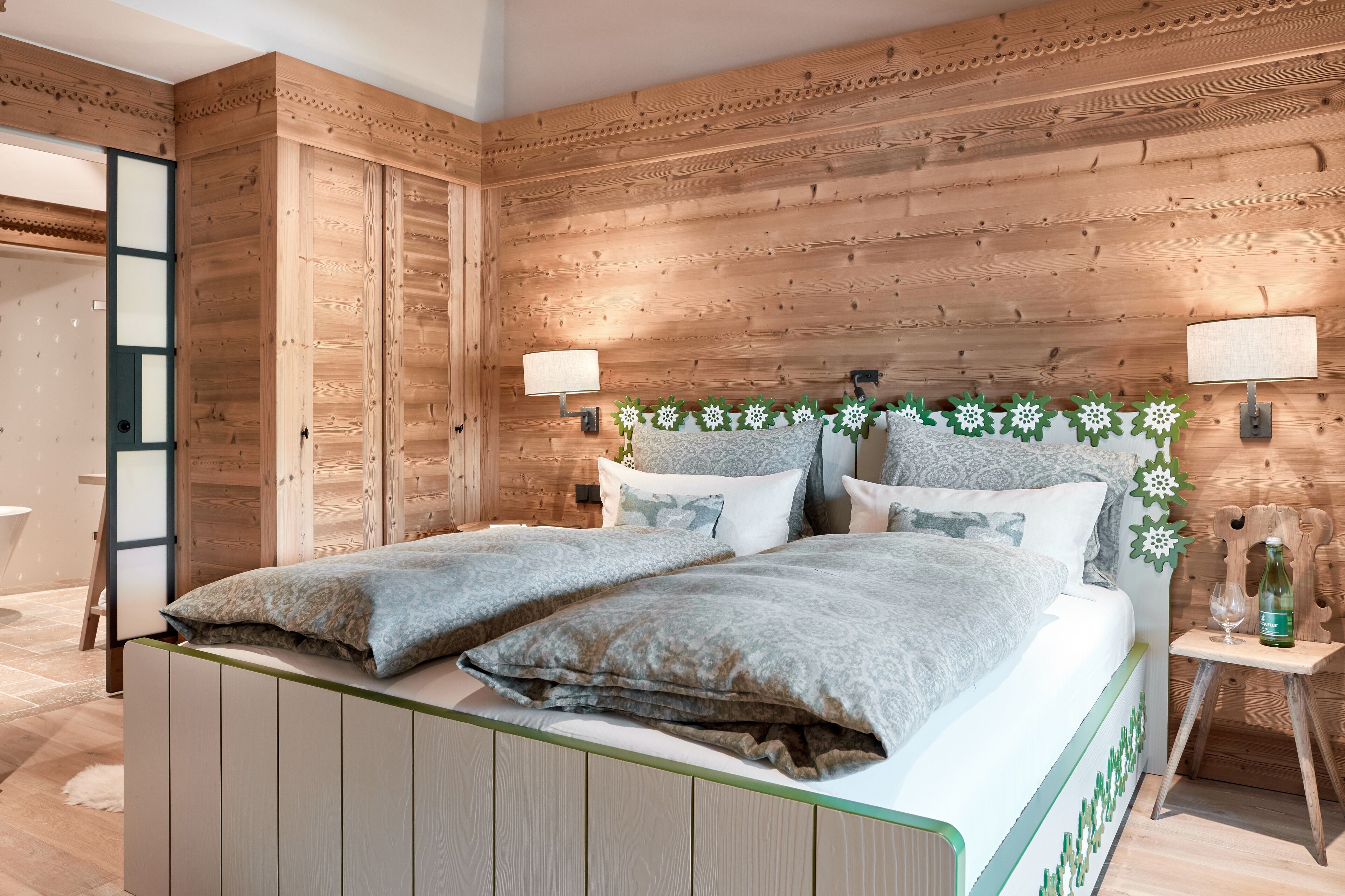 A wooden-paneled bedroom with a bed, two bedside lamps, and a small table with a bottle and glass.