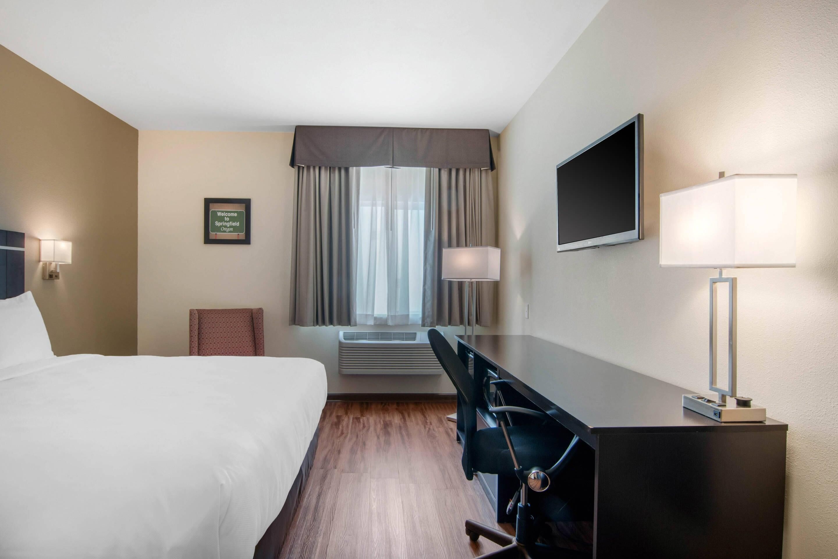 Habitación de hotel con cama, escritorio, silla, televisor, lámpara, equipo de aire acondicionado y un cartel de bienvenida enmarcado.