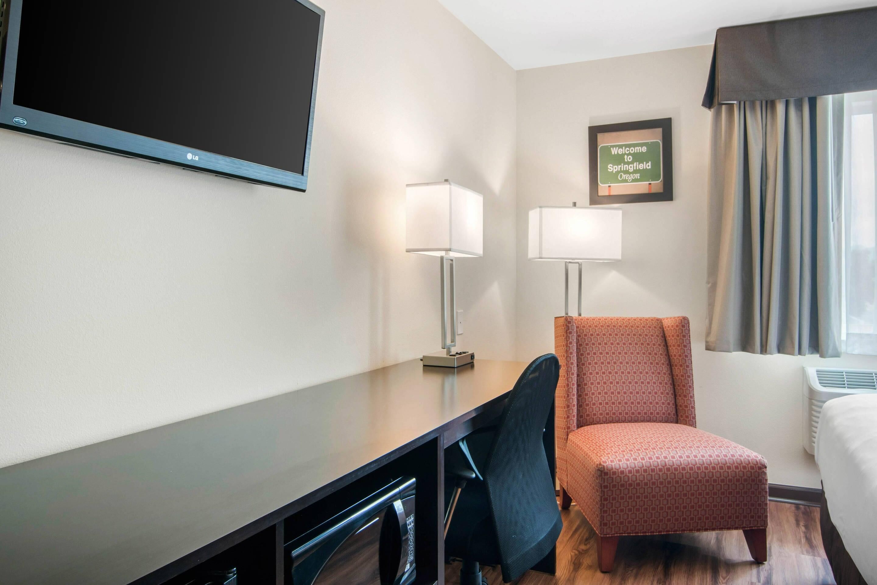 A hotel room with a flat-screen TV, two bedside lamps, a framed welcome sign, a window with curtains, and a wooden desk with a chair.