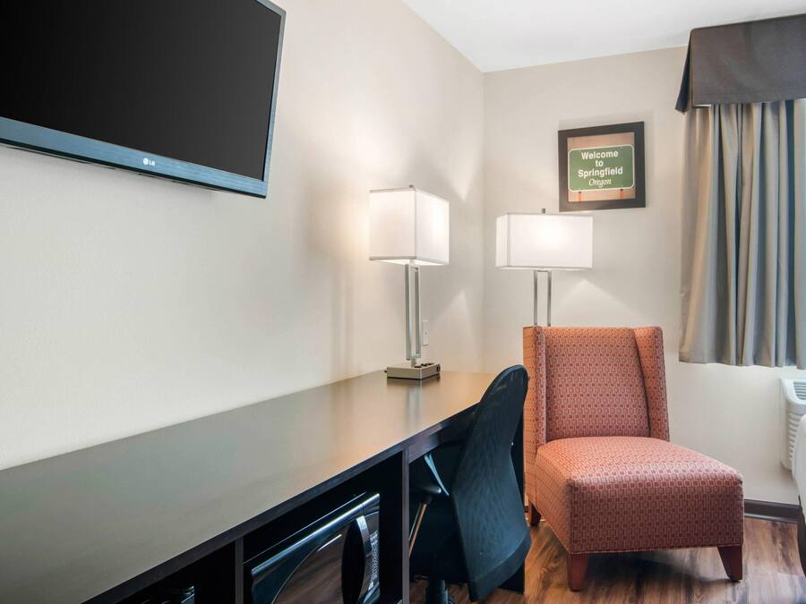 A hotel room with a flat-screen TV, two bedside lamps, a framed welcome sign, a window with curtains, and a wooden desk with a chair.