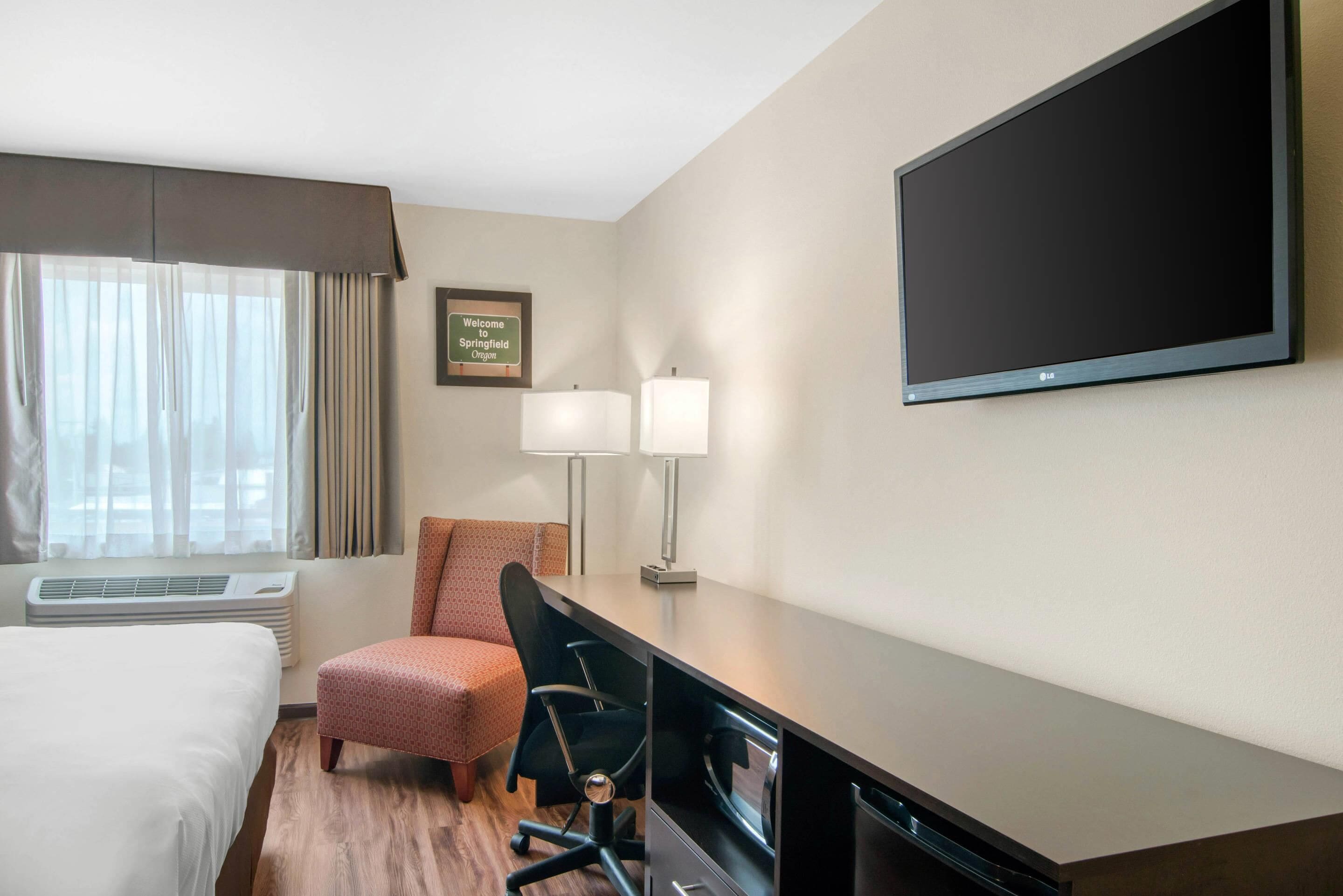 A hotel room with a bed, a desk, a chair, a TV, a lamp, and a sign on the wall.