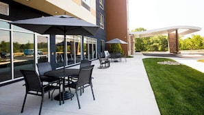 An outdoor patio area with tables and chairs, large umbrellas, and a building with large windows and a stone facade. - Fairfield Inn & Suites Philadelphia Broomall/Newtown Square (Broomall)