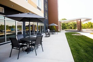 Un patio al aire libre con mesas y sillas, sombrillas grandes, y un edificio con ventanas amplias y fachada de piedra.