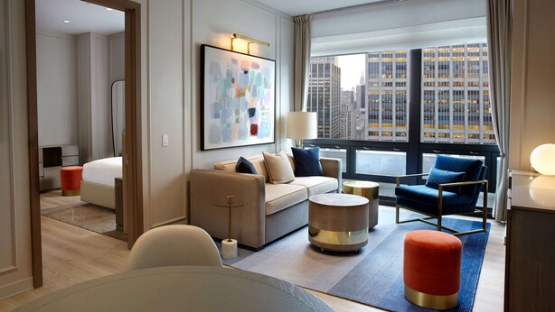 A modern hotel room with a large window offering a city view, a comfortable seating area with a sofa and chairs, a small round table, and a bed with a red ottoman. - The Residences by Hilton Club (New York)