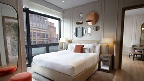 A modern hotel room with a large bed, a sitting area with a chair, a desk, and a large window offering a city view. - The Residences by Hilton Club (New York)