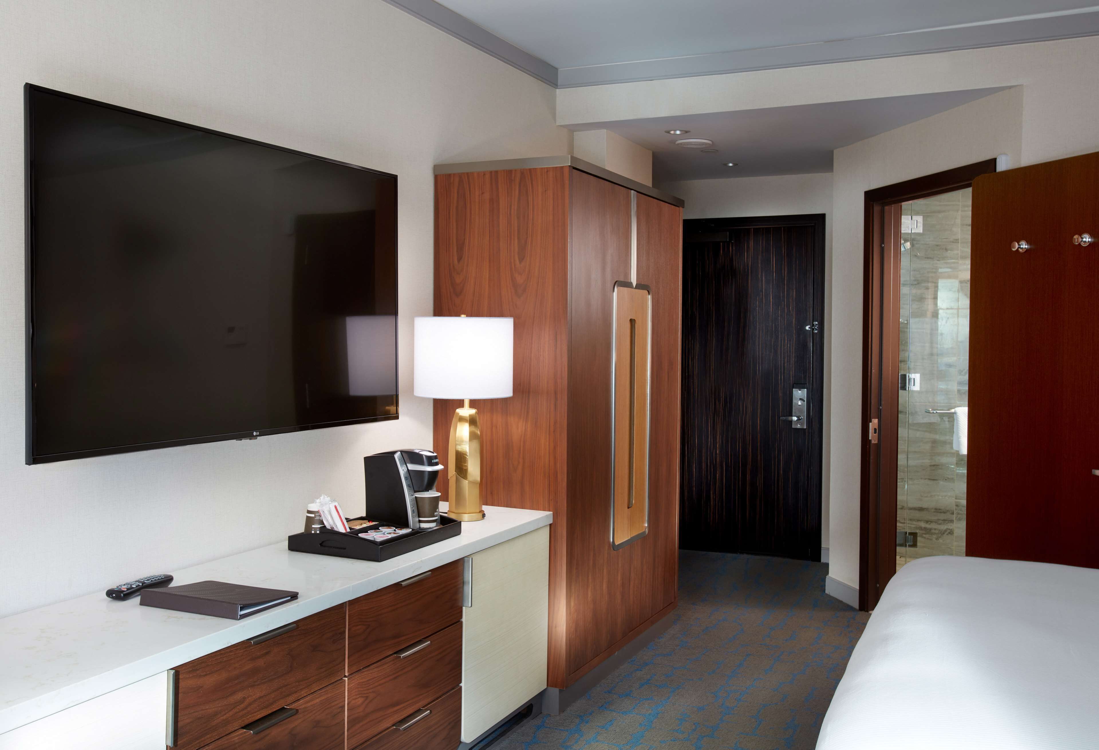 A hotel room with a flat-screen TV, a desk with a lamp, a minibar, a wardrobe, and a bed.