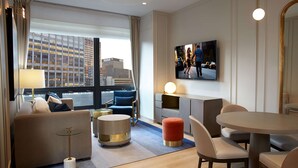 A modern hotel room with a large window, a flat-screen TV, a sofa, a round coffee table, and a dining area with a table and chairs. - The Residences by Hilton Club (New York)