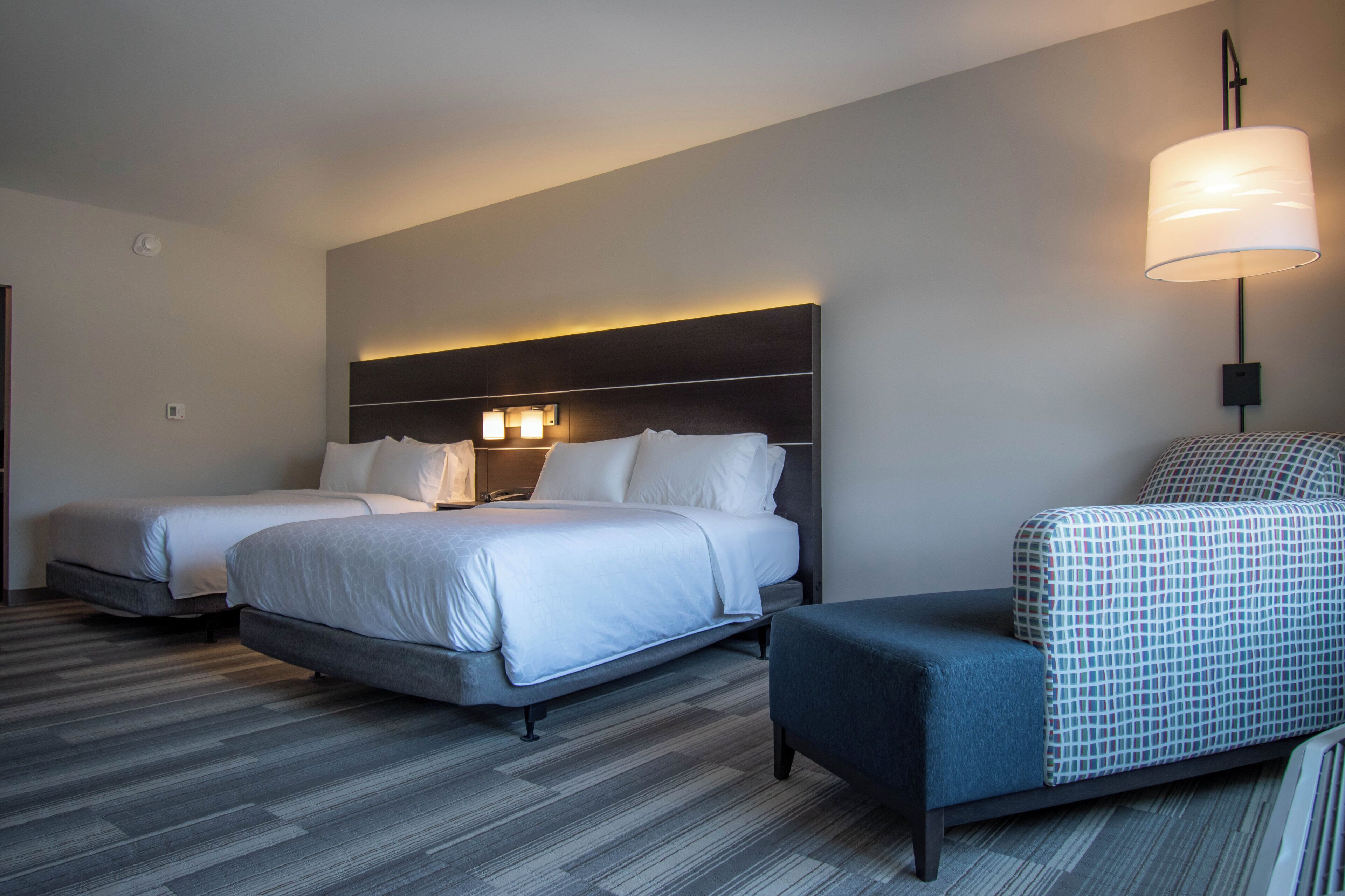 A hotel room with two beds, a desk, a chair, a lamp, and a patterned ottoman.