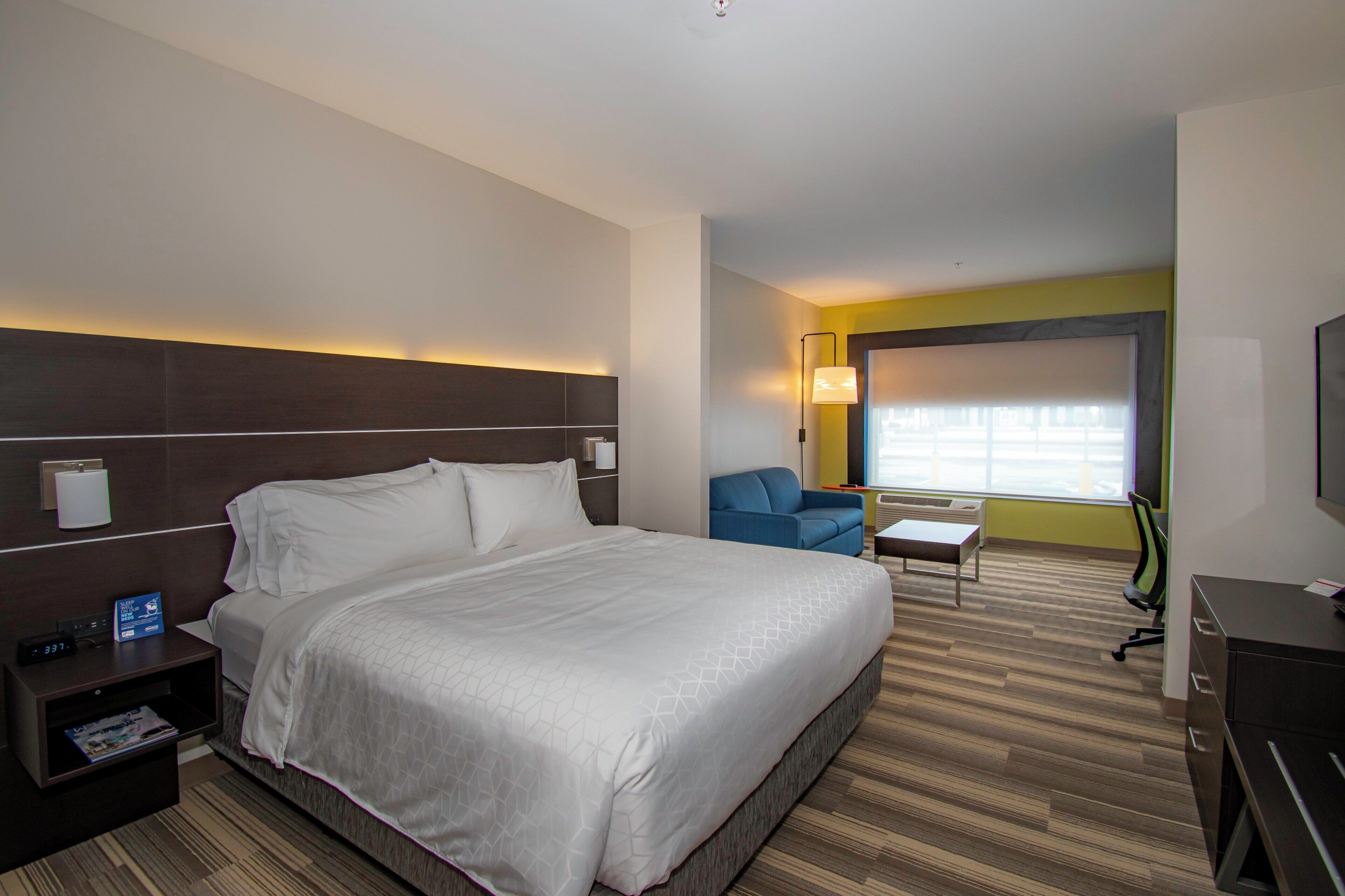 A hotel room with a large bed, a desk, a chair, a TV, and a window with blinds.