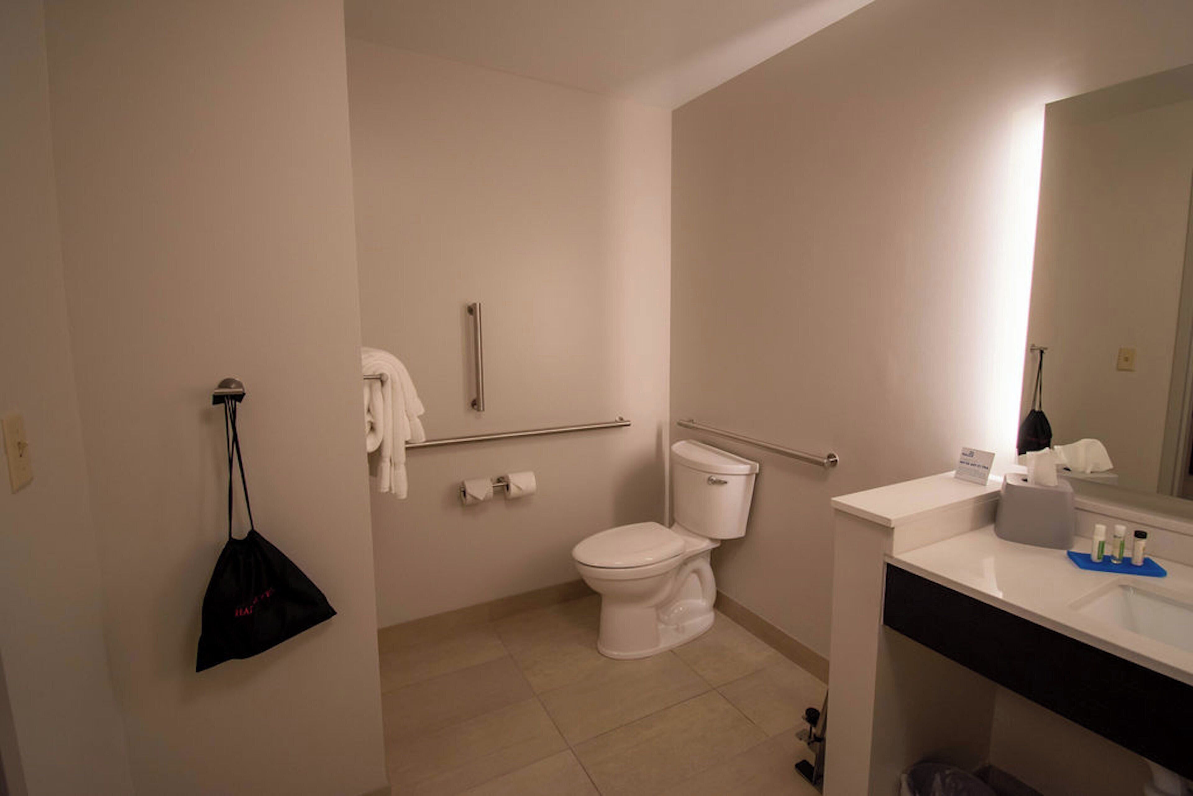 A standard hotel bathroom with a toilet, handrail, towel rack, and a sink with toiletries.