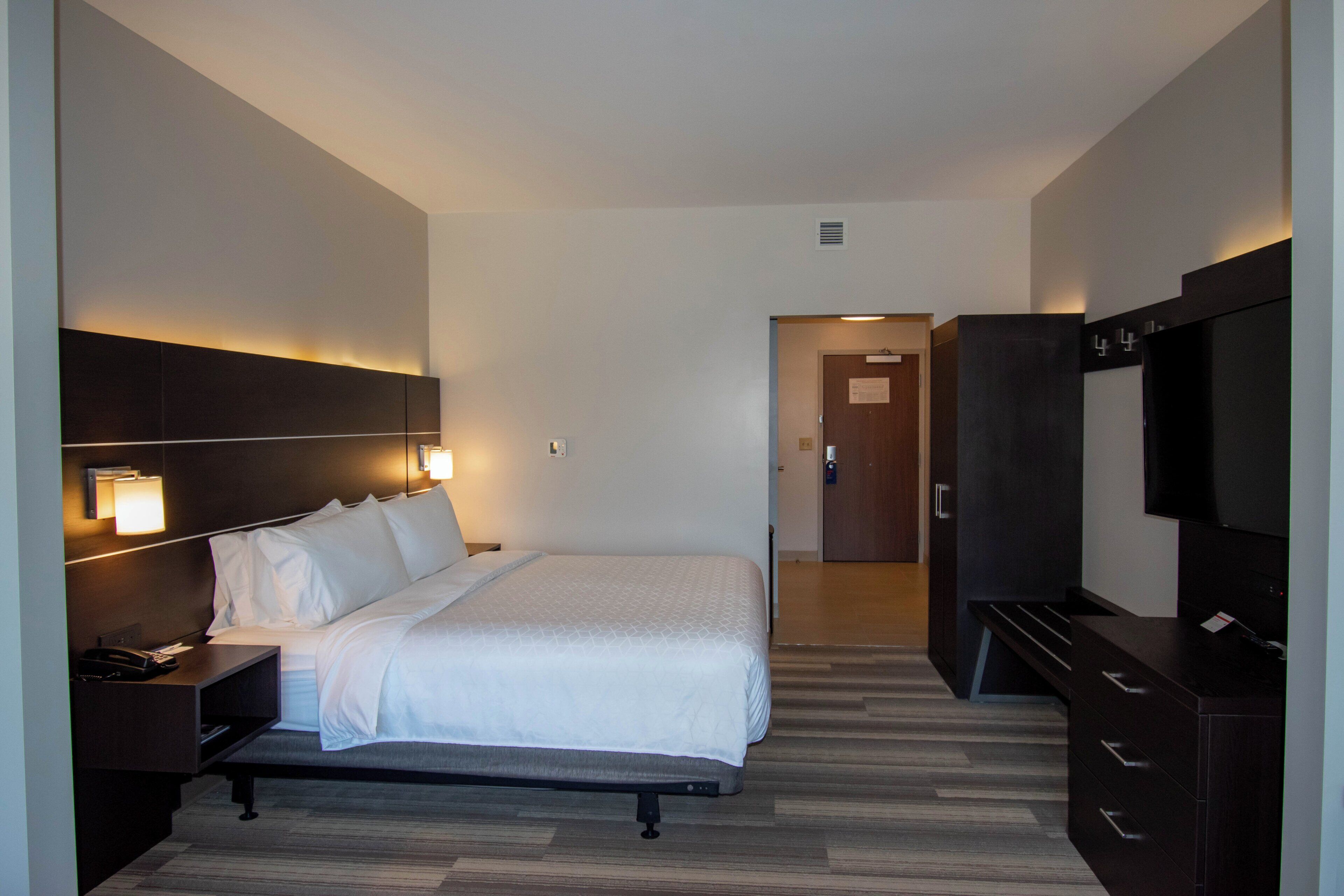 A hotel room with a bed, bedside table, lamp, TV, wardrobe, and a door leading to another room.