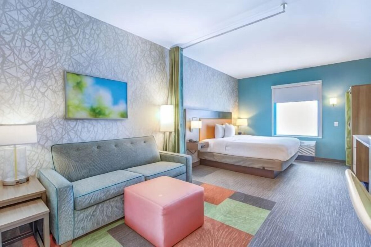 Une chambre d’hôtel avec un lit, un canapé, un pouf rose, un bureau avec une lampe et un tableau au mur.