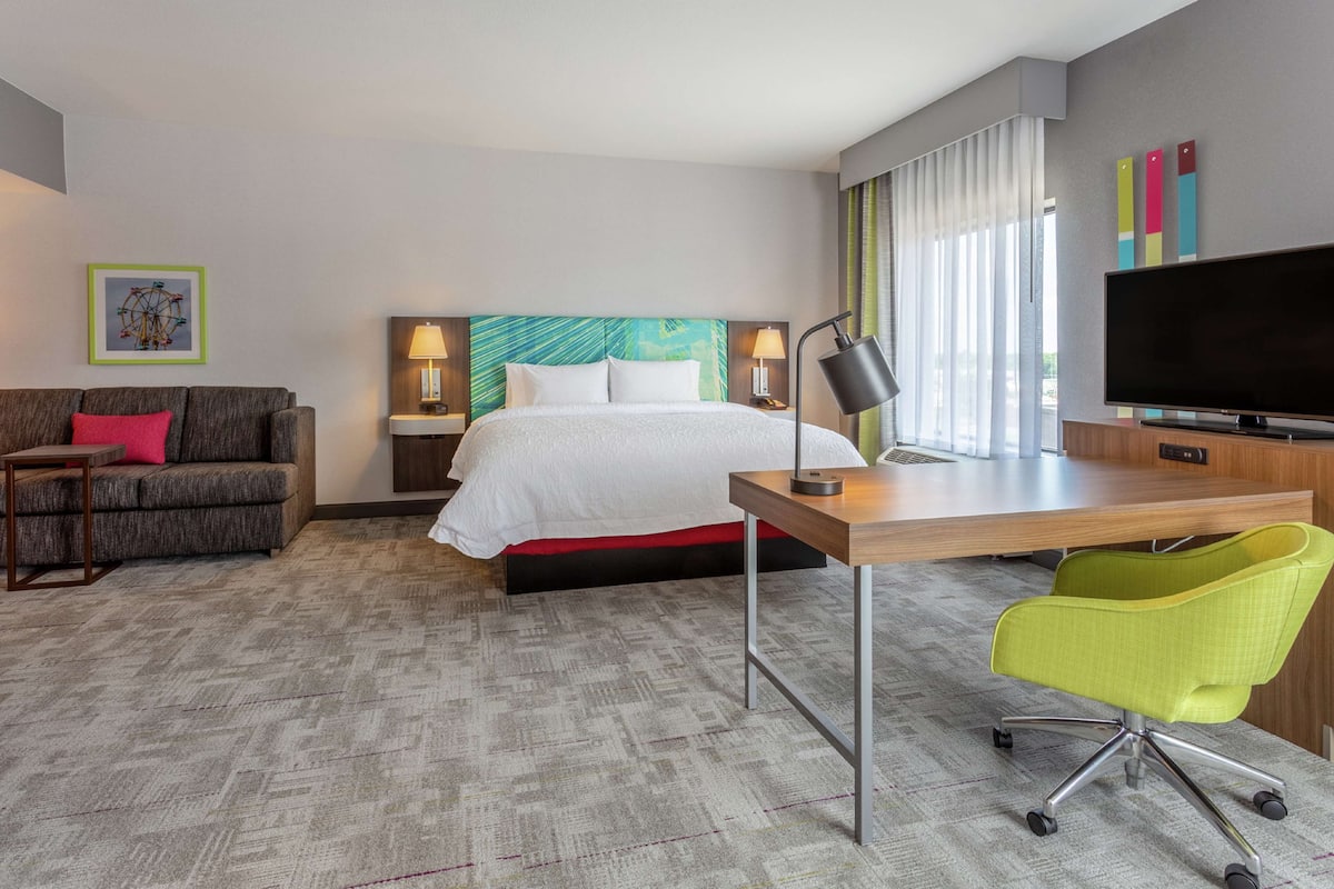 Une chambre d’hôtel avec un lit, un bureau, une chaise, une télévision, une lampe, un canapé et un tableau au mur.