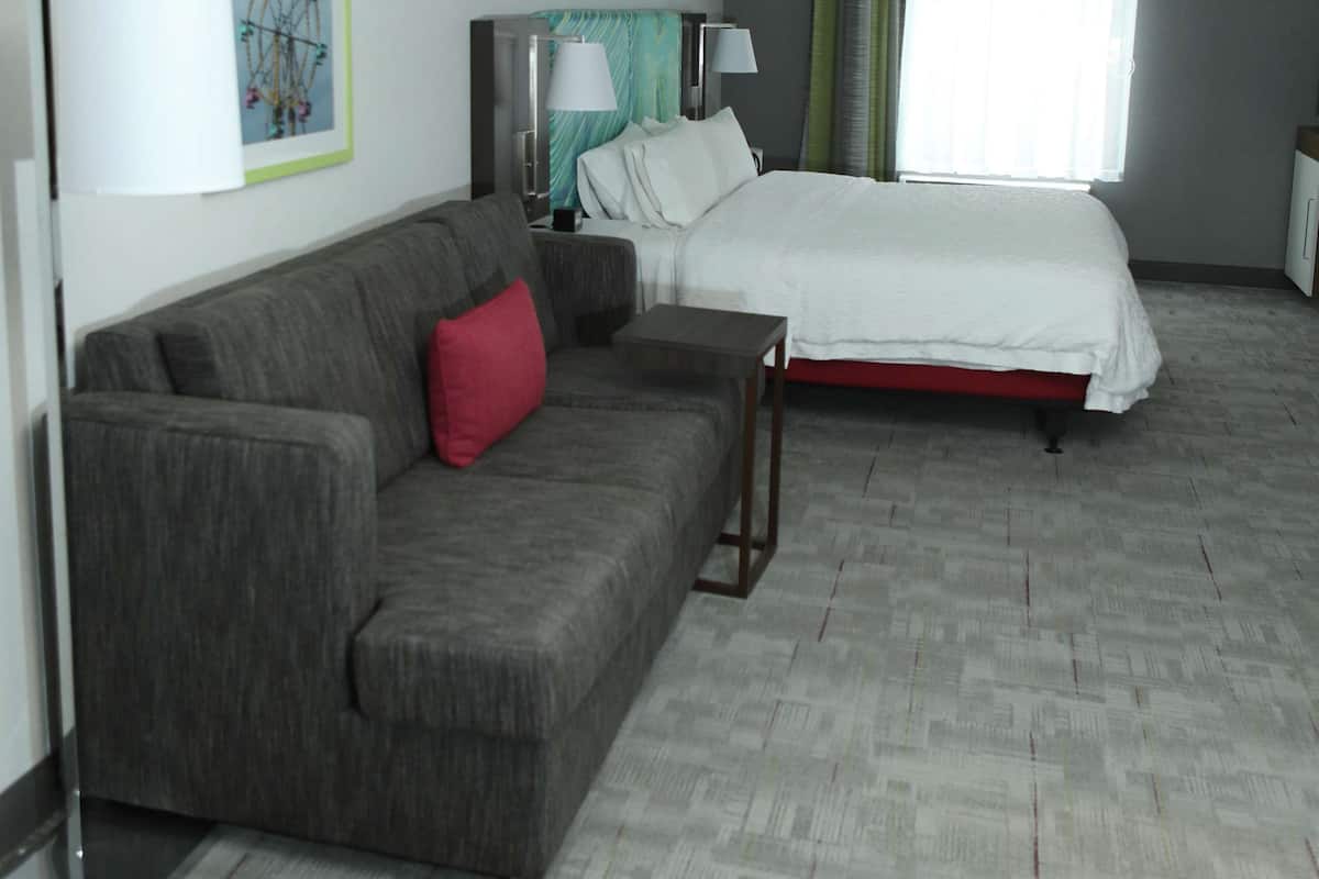 Une chambre d’hôtel avec un canapé gris, un lit avec une literie blanche, un bureau et une fenêtre avec des rideaux.