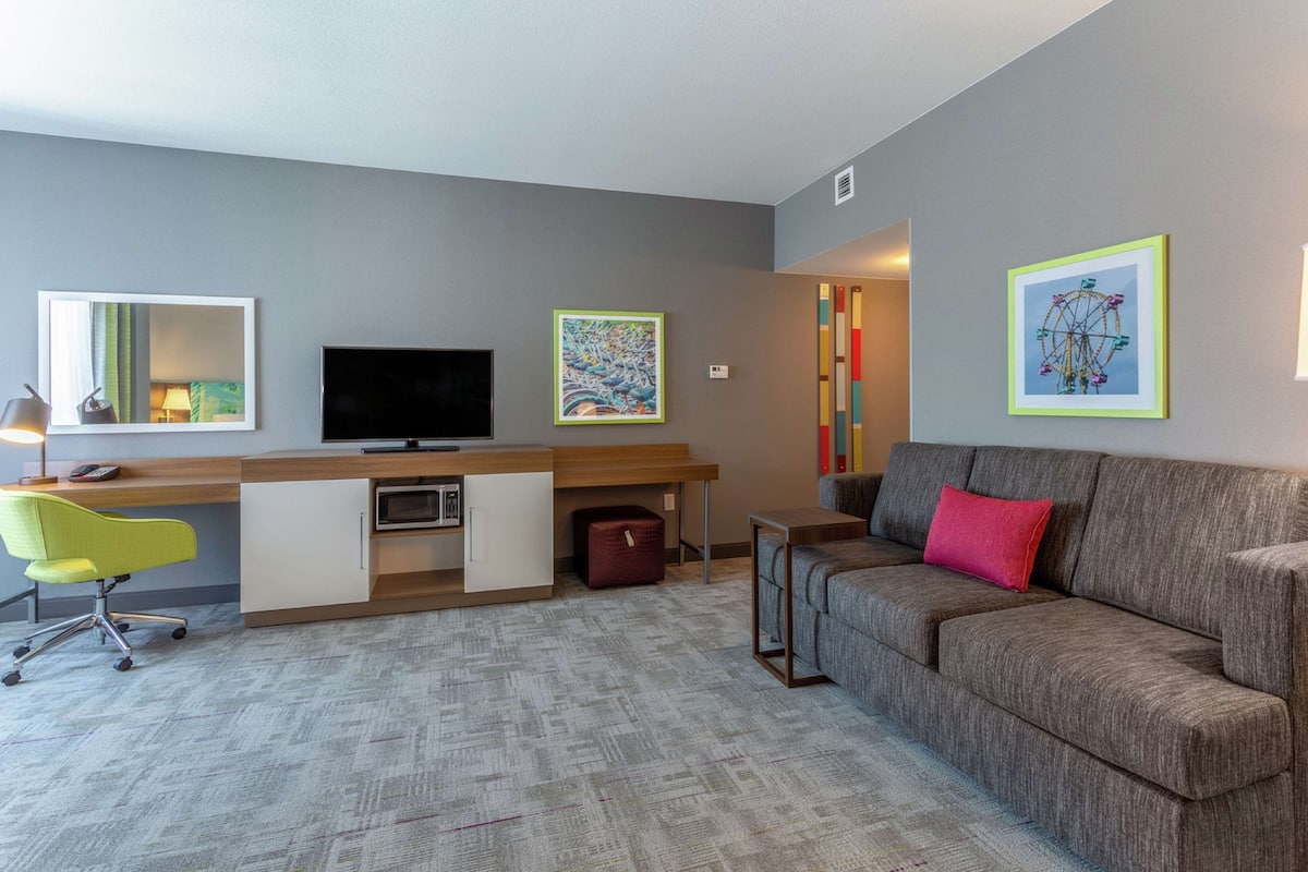 Une chambre d’hôtel moderne avec un canapé gris, un bureau avec une chaise, une télévision et des œuvres d’art accrochées aux murs.