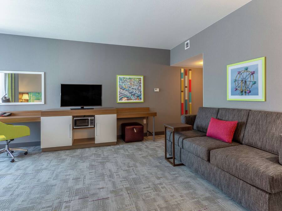 A modern hotel room with a grey sofa, a desk with a chair, a television, and artwork on the walls.