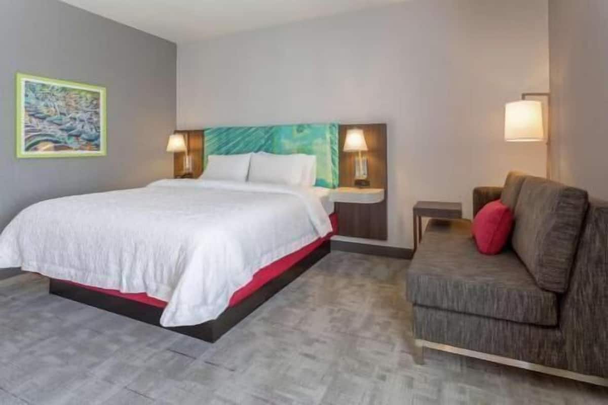 A hotel room with a bed, a sofa, a nightstand, a lamp, and a painting on the wall.