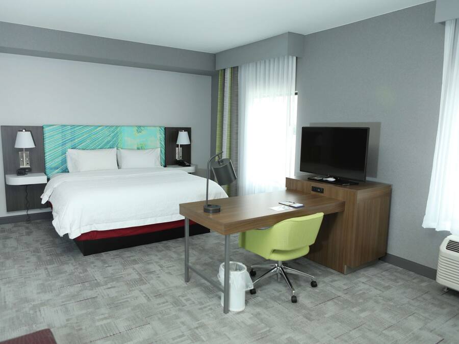 A hotel room with a bed, desk, chair, TV, and a waste bin.